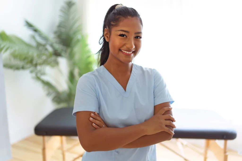 A female massage therapist wearing light blue scrubs smiles and crosses her arms while standing in front of a black massage table.