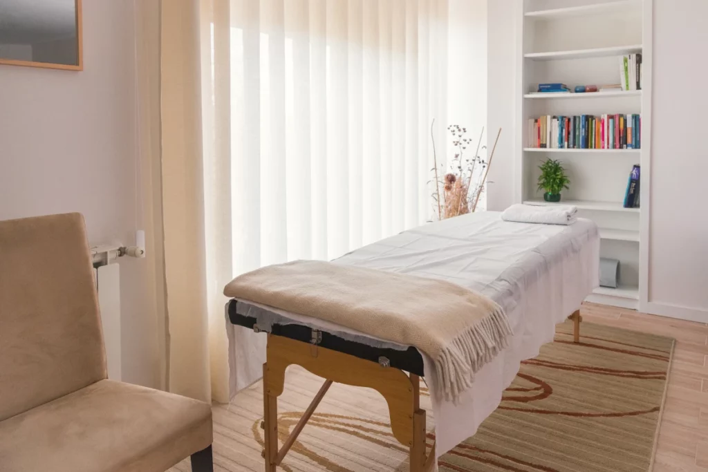 A massage room with a massage table draped in white sheets, white curtains, a white bookshelf with colorful books, and a beige chair.