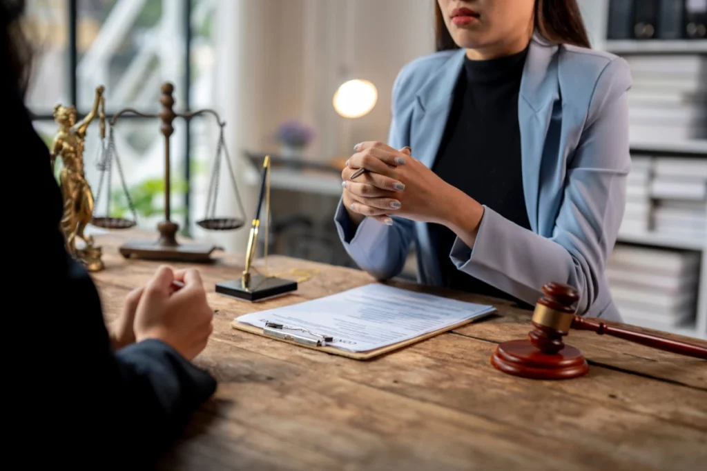 A lawyer dressed in a black top and a blue blazer sits across a client with a clipboard, pen, gavel, and justice symbols on the wooden desktop.