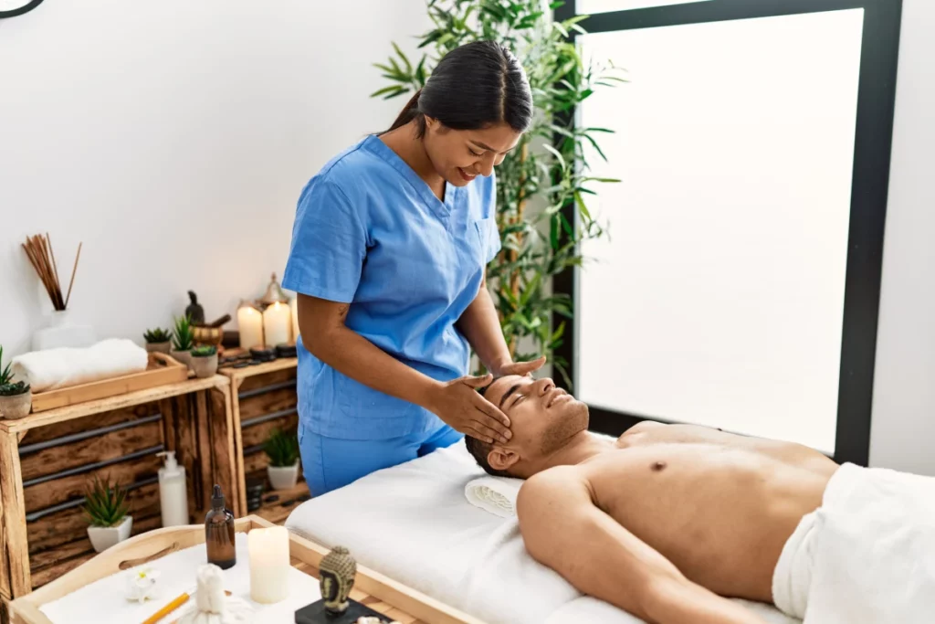 A female massage therapist wearing blue scrubs massages the temples of a male patient who is lying on a massage table.