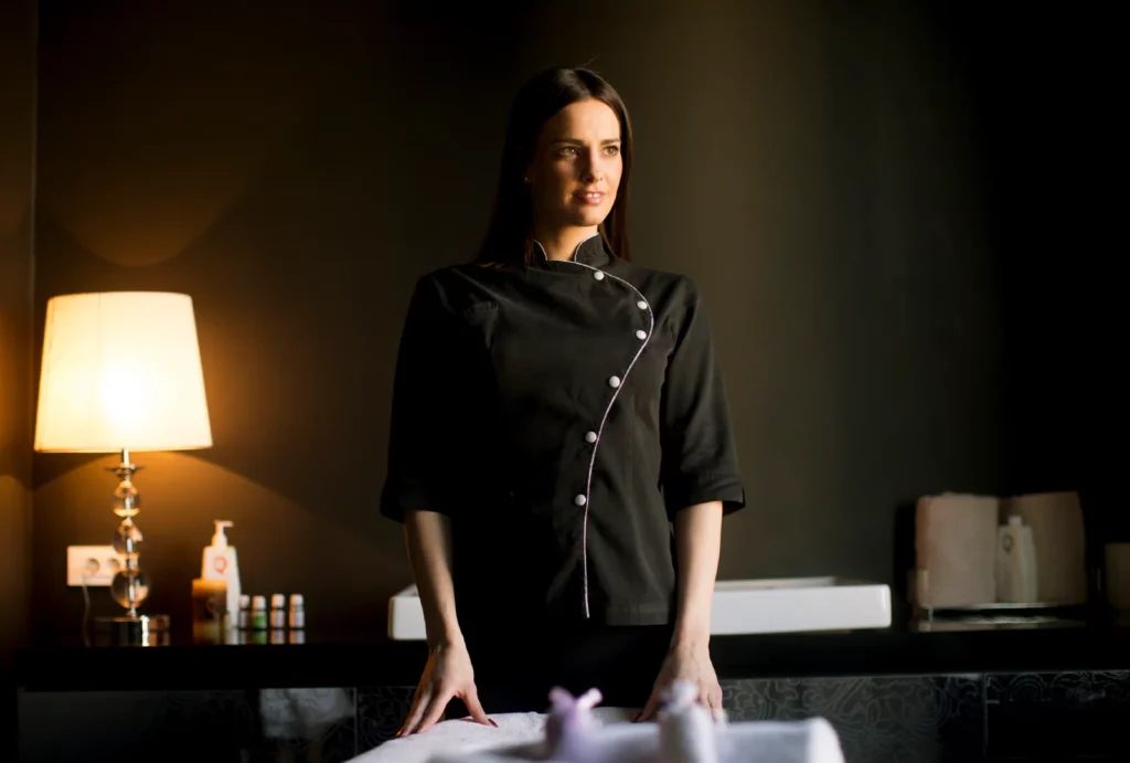 A female massage therapist wearing a black uniform smiles and looks into the distance in her massage room.