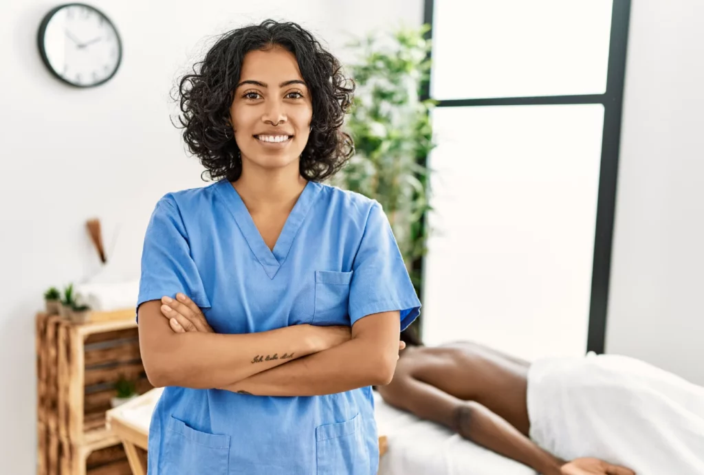 A massage therapist with curly hair wearing blue scrubs smiles and poses with her arms crossed in front of her massage station. In the background, a client lies on the massage table with a white towel on their body.