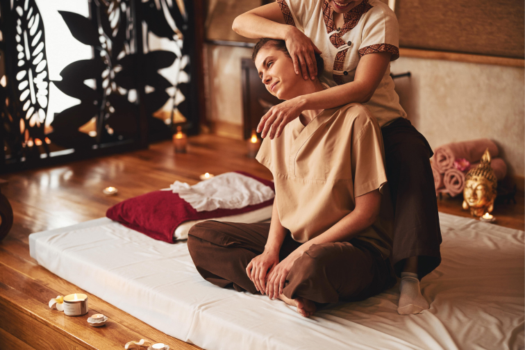 A Thai massage therapist stretches the neck of a female patient in a wooden massage room with a white massage mat and candles.