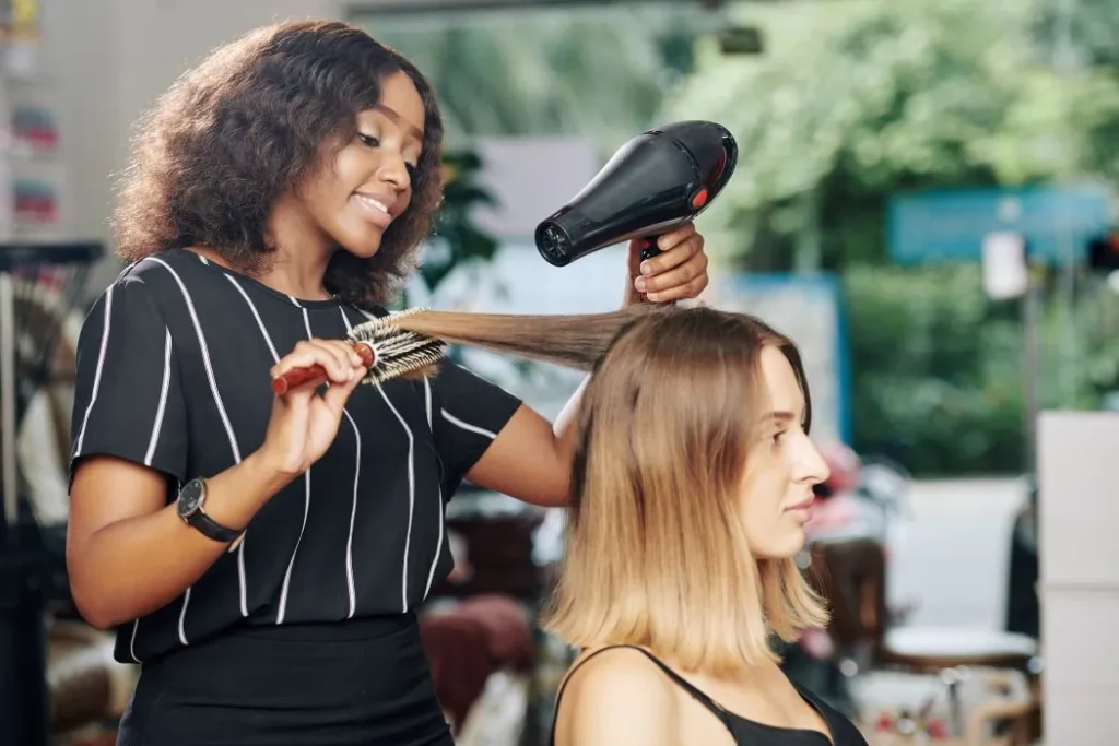 A female hair stylist smiles while blow-drying her client's hair with a round brush.