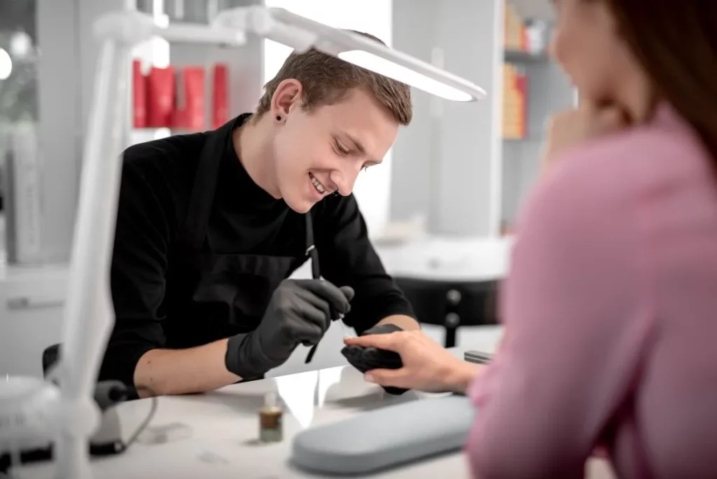 Young male manicurist smiles while painting client's nails.