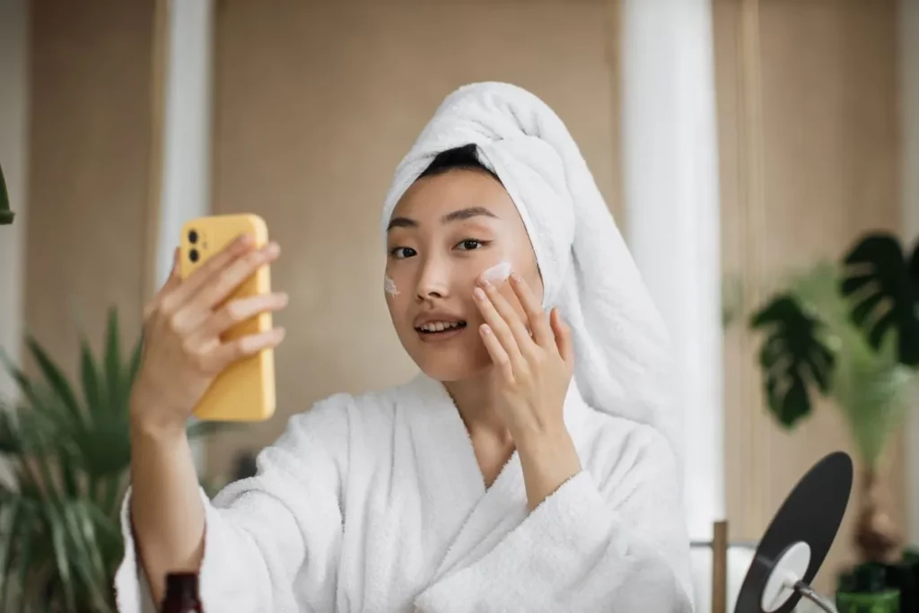 Female esthetician wearing a towel on her hair applies moisturizer to her cheek while filming with a smartphone.
