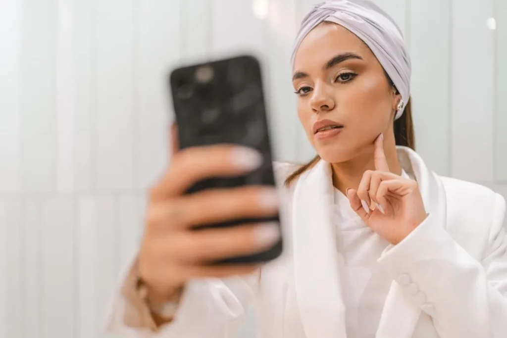 Esthetician wearing a white robe checking her jawline while looking into a black smartphone.