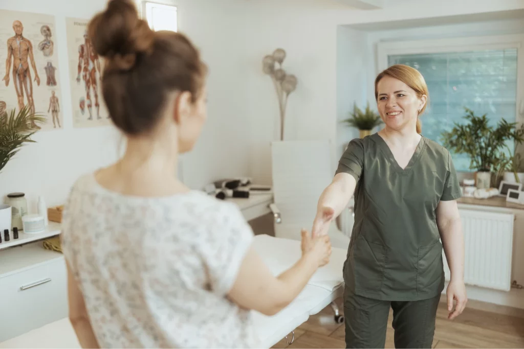 A female massage therapist wearing green scrubs smiles and shakes hands with a client in a massage studio.