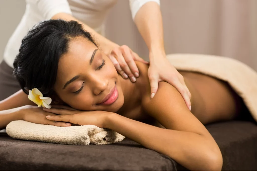 A woman wearing a plumeria flower smiles while receiving a shoulder massage from a massage therapist.