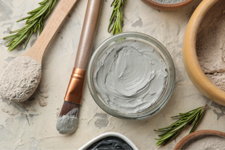 Clay mask in a jar surrounded by various tools, rosemary herbs, and bowls of dry clay mixture for estheticians.