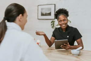 Female esthetician wearing a black uniform and holding a tablet explains treatment to a client with a ponytail in a white robe at a wooden table.