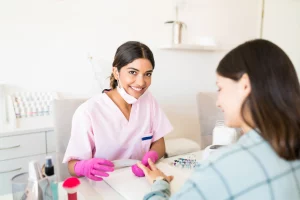 A young manicurist wearing pink gloves files a client's nails at her nail salon station.