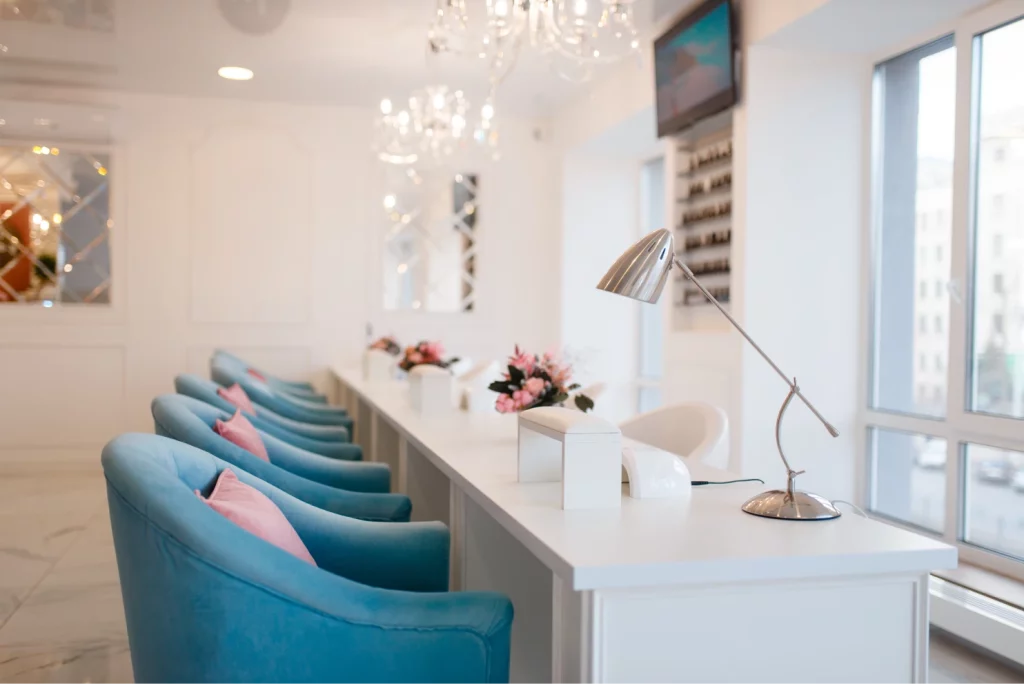 A nail salon with a minimalist white interior, plush blue chairs, LED curing lamps on the tables, and chandeliers on the ceiling with a city view from the window.