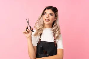 A blonde hair stylist wearing a white top and an apron holds a pair of scissors and smiles while looking off to the side against a pink backdrop.