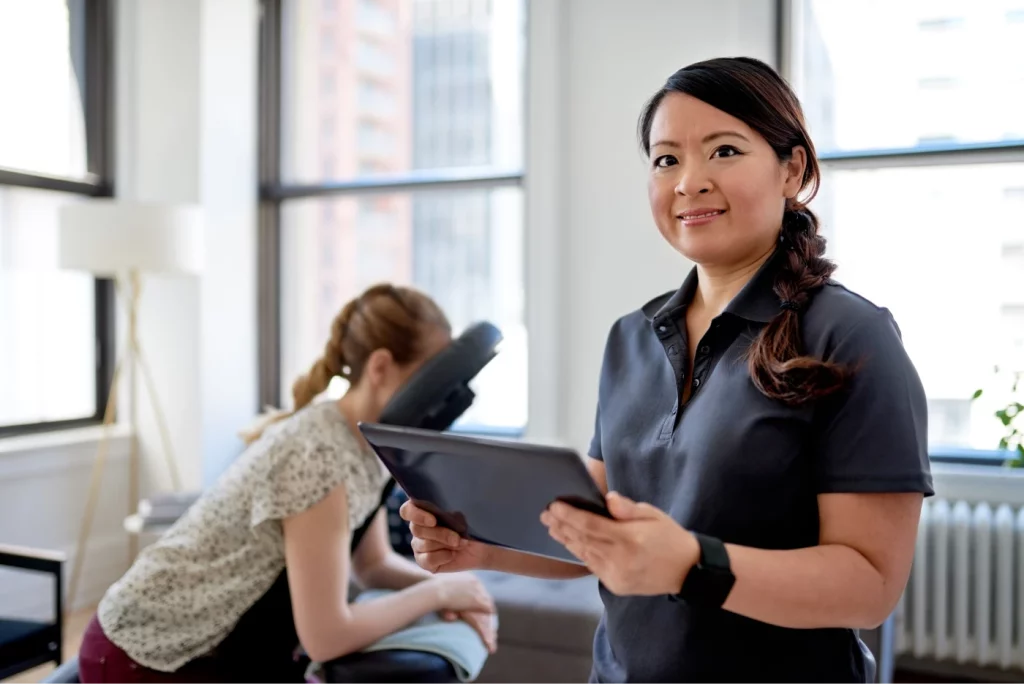 A massage therapist wearing a black polo top smiles and holds a tablet while her client sits at a massage chair in the background of her workspace.