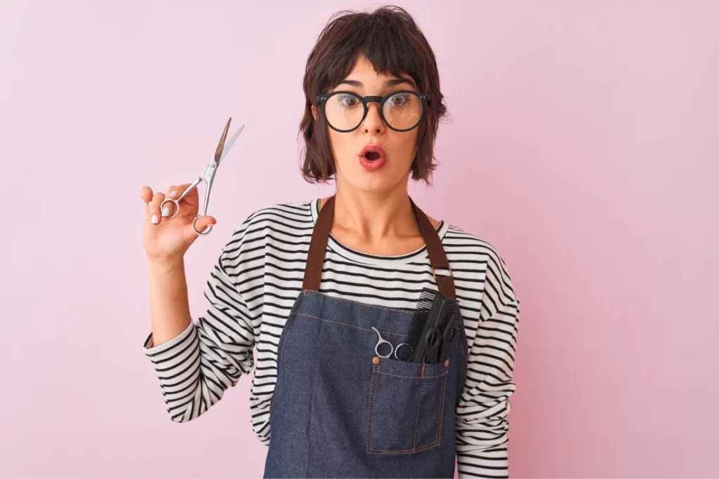 A shocked hair stylist with short hair and glasses wearing a striped top and an apron holds a pair of scissors while standing against a pink backdrop.