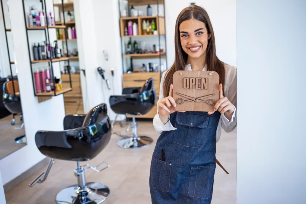 A brunette hair stylist wearing a denim apron smiles and holds a wooden sign that says "open" in a hair salon.