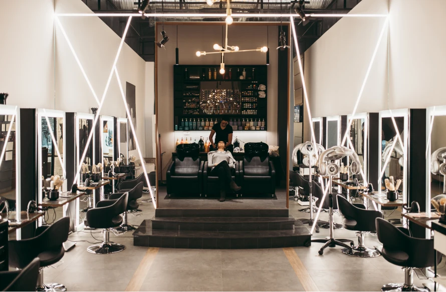 A modern hair salon with rows of stations on either side, bright tube lighting, and a shampoo station in the middle. A stylist shampoos a client's hair in the middle shampoo chair.