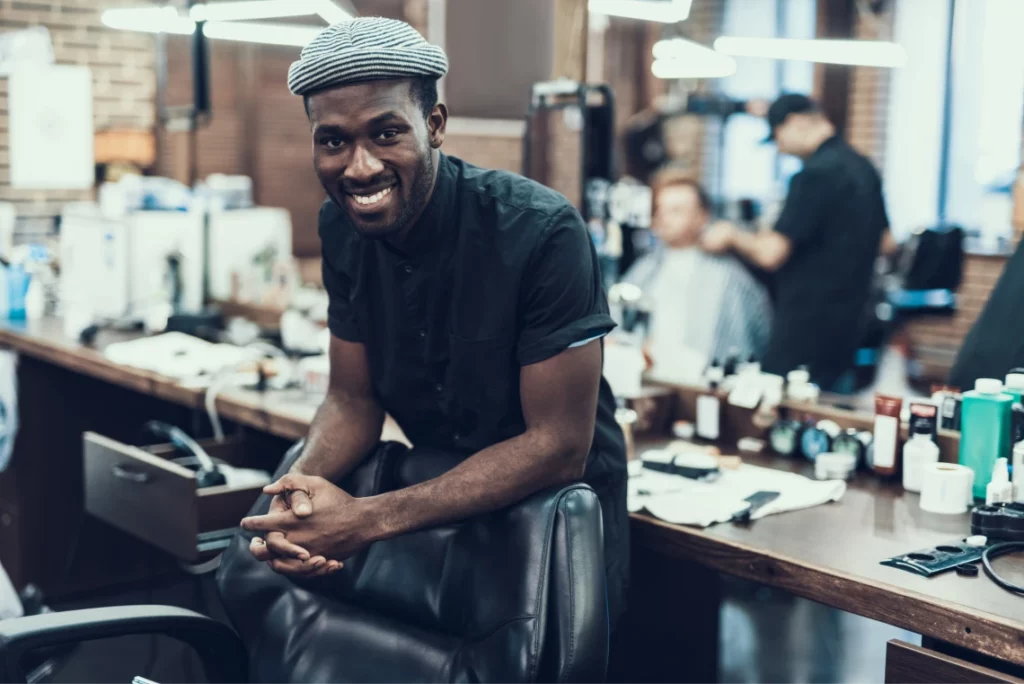 A young male barber wearing a striped beanie and a black uniform rests his forearms on a barber chair and smiles in a barbershop.