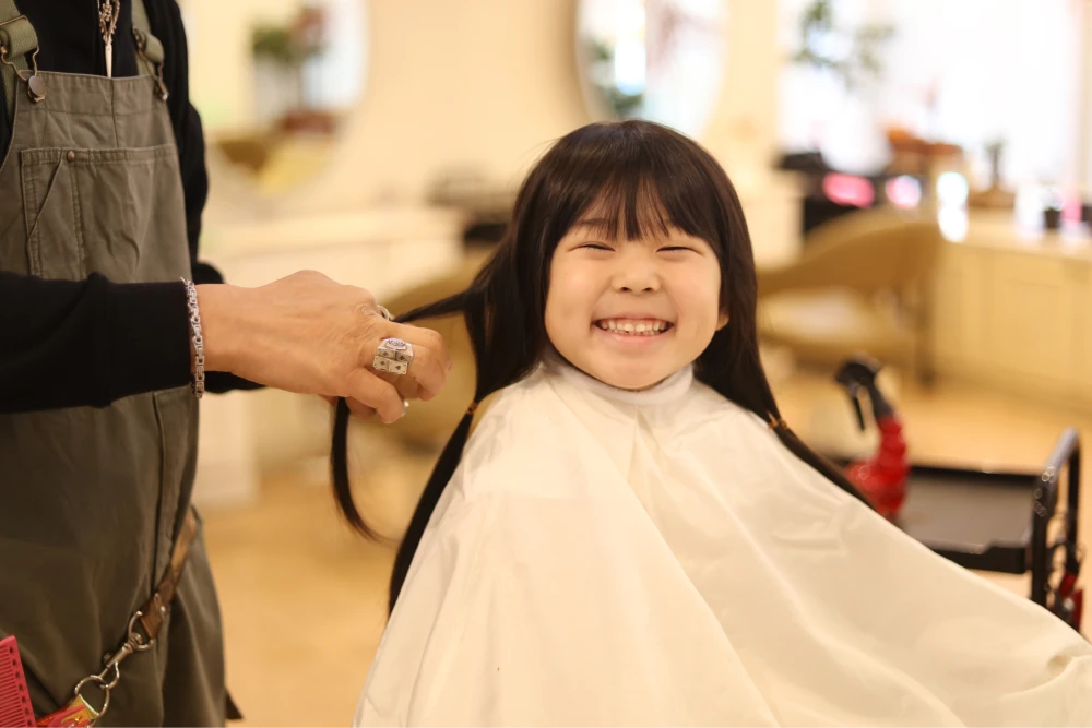 A young girl with straight black hair and bangs wearing a white apron smiles while having her hair done by a stylist in a salon.