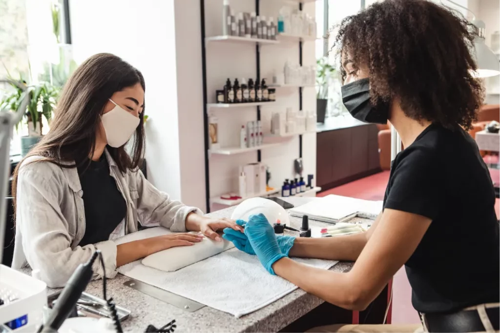 A nail tech wearing a face mask applies light pink gel nail polish to a client's nails in a nail salon.