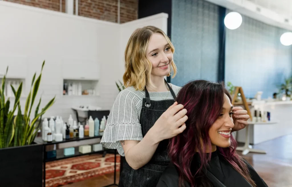 A blonde hair stylist wearing a denim apron fixes her client's burgundy hair in waves in a bright salon.