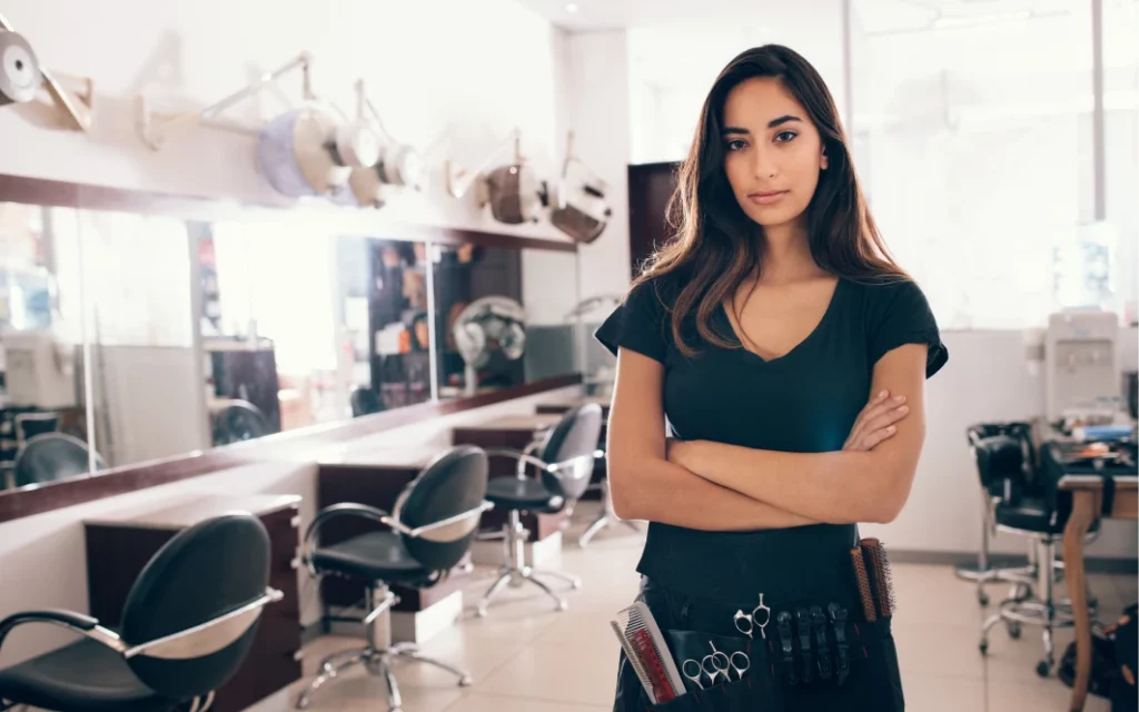 A female hair stylist wearing a black shirt and stylist apron poses with her arms crossed in a hair salon.