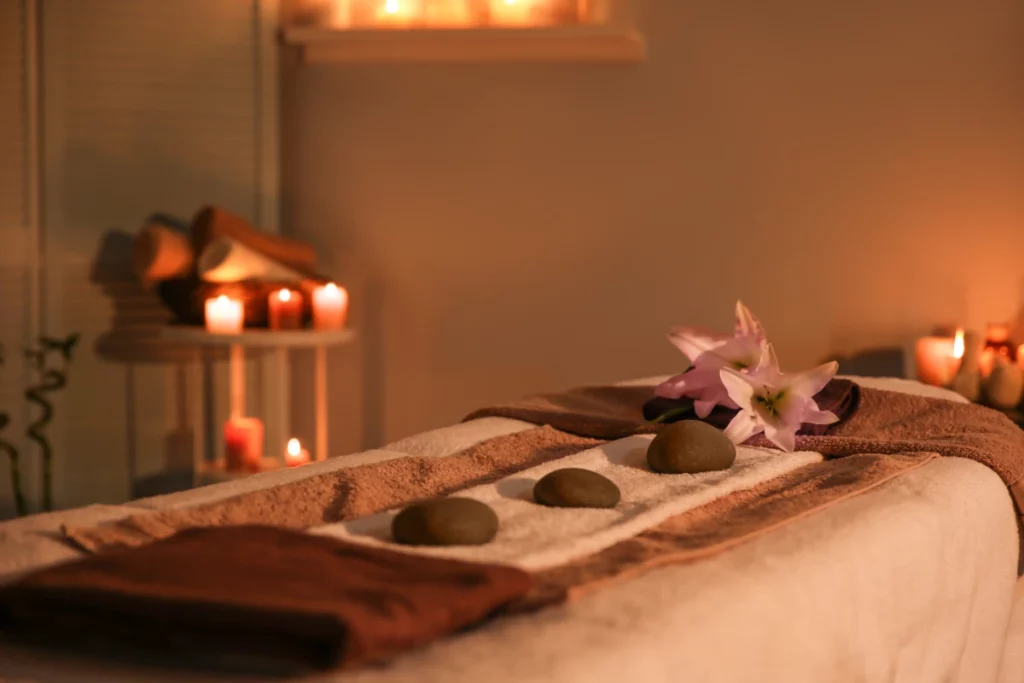 A massage table set with white and brown towels, gray stones, and purple flowers in a candlelit massage room.